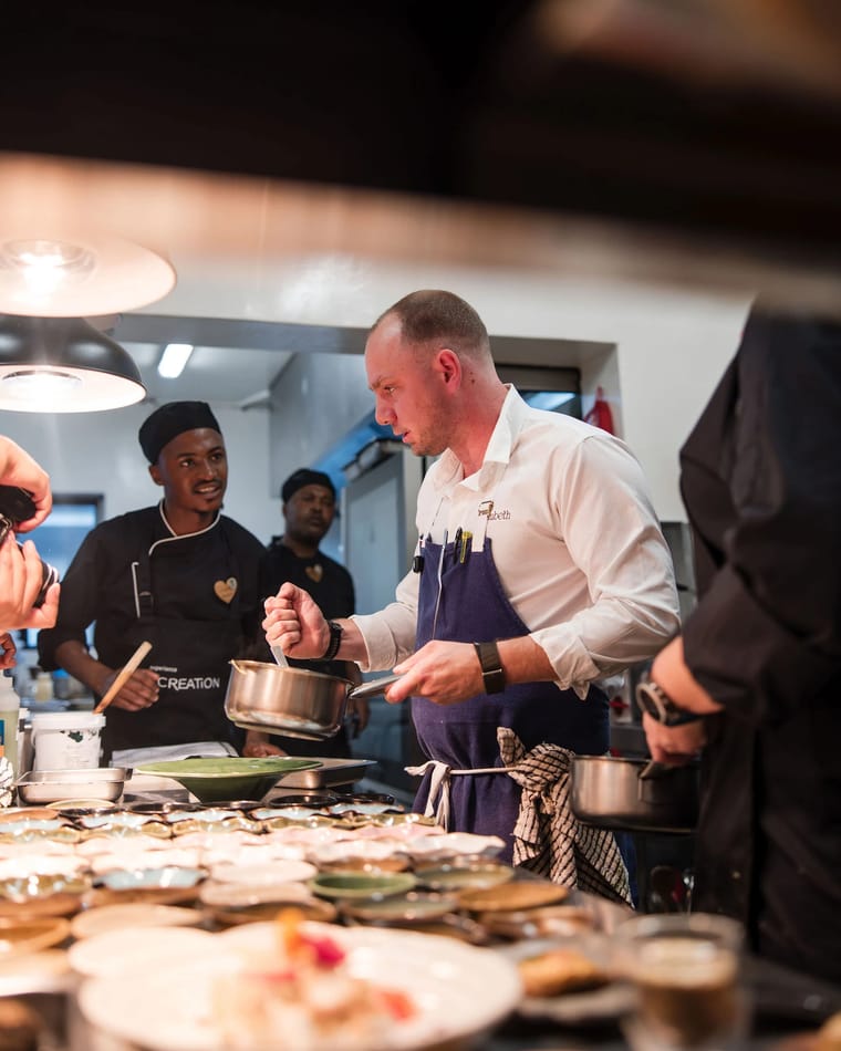 Chef Jason McKinney holding a saucepan over a pass full of plates, with the Creation Wines kitchen team behind him